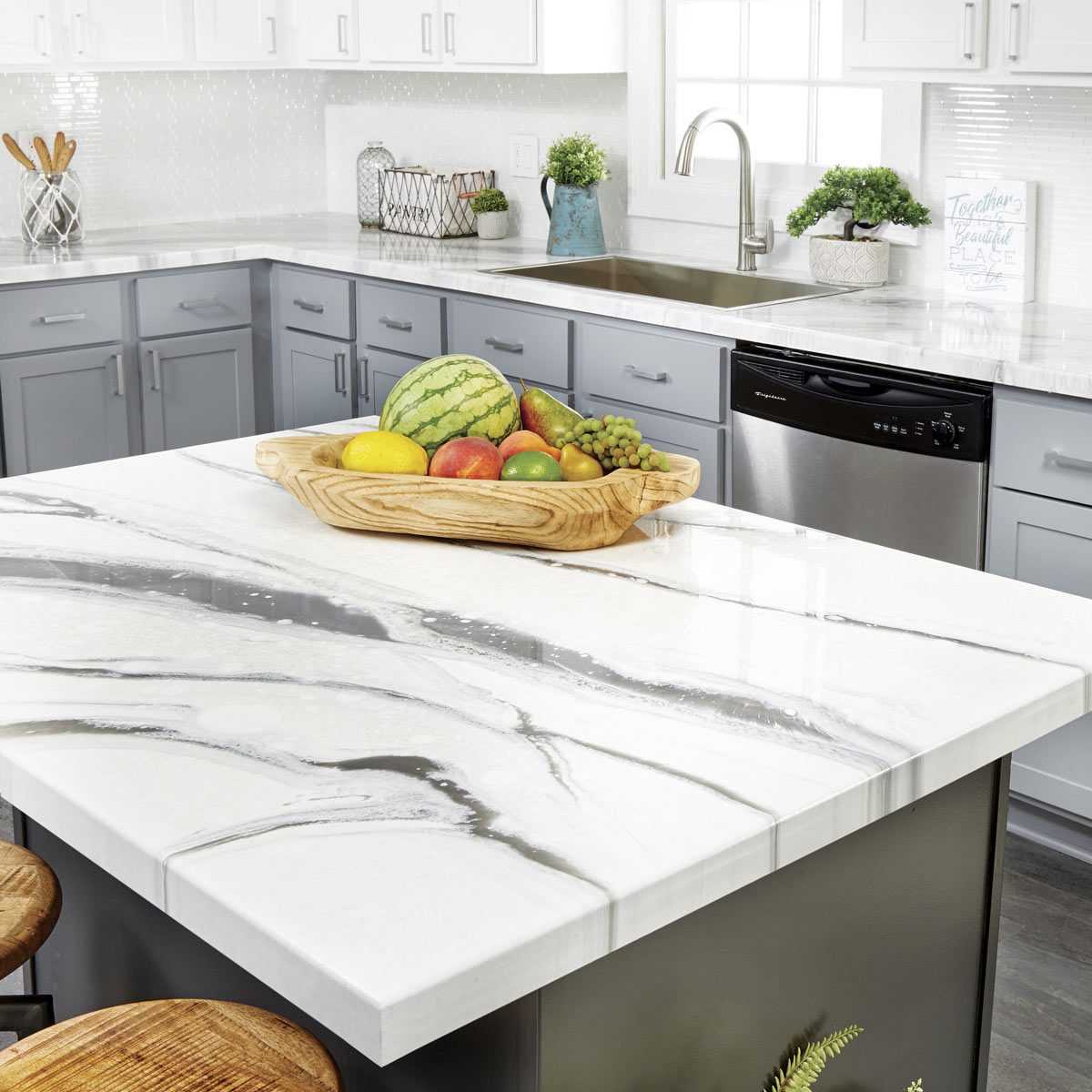 A wooden bowl filled with various fruits sits on a shiny, marble-patterned kitchen countertop, surrounded by gray cabinets and a stainless steel sink.
