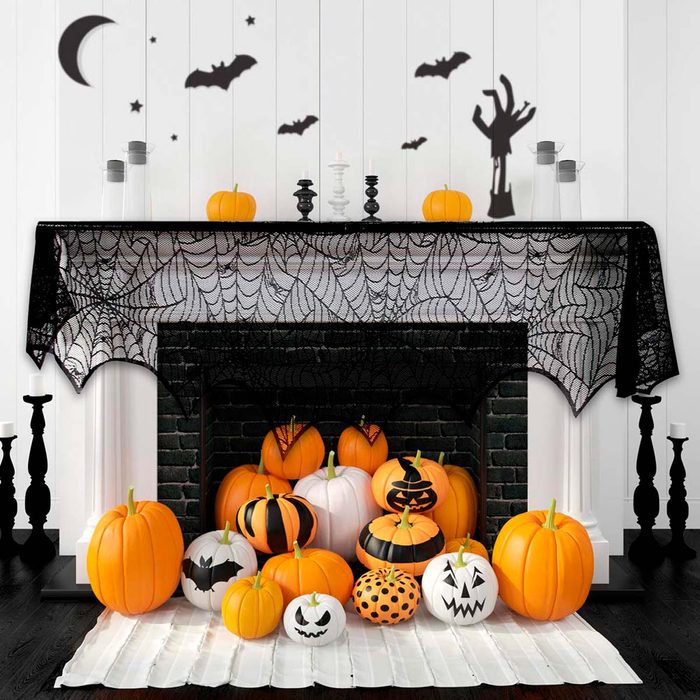 A collection of decorated pumpkins sits on a white mat by a black fireplace, surrounded by Halloween decorations like bats and spider webs on the wall.