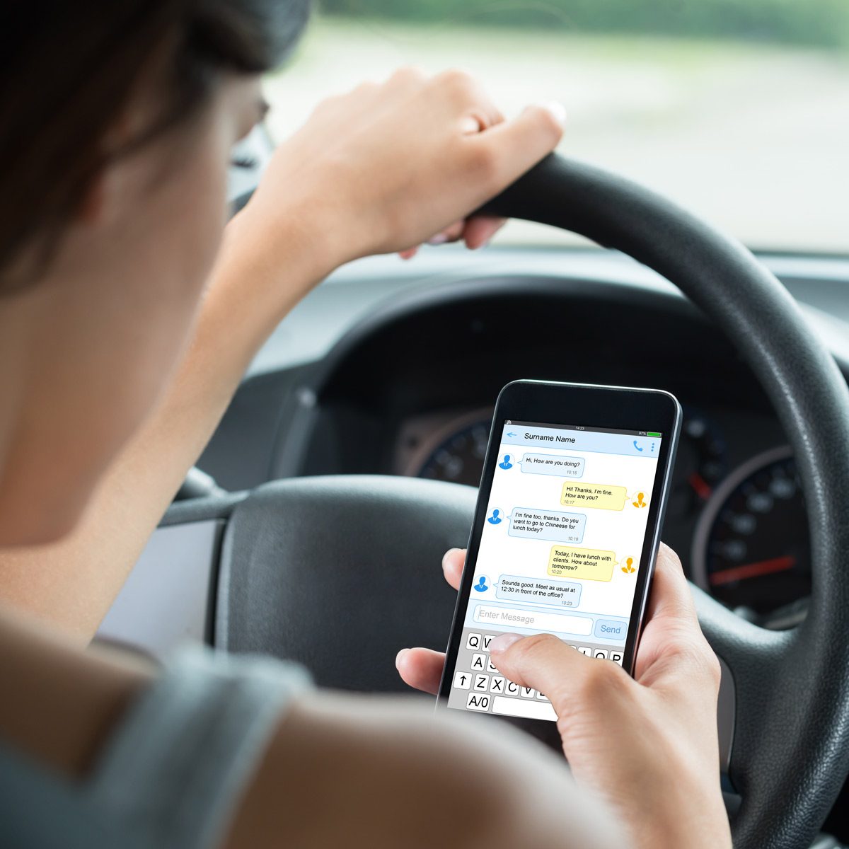 A person is texting on a smartphone while gripping the steering wheel of a car, focused on the screen as the vehicle is in motion.
