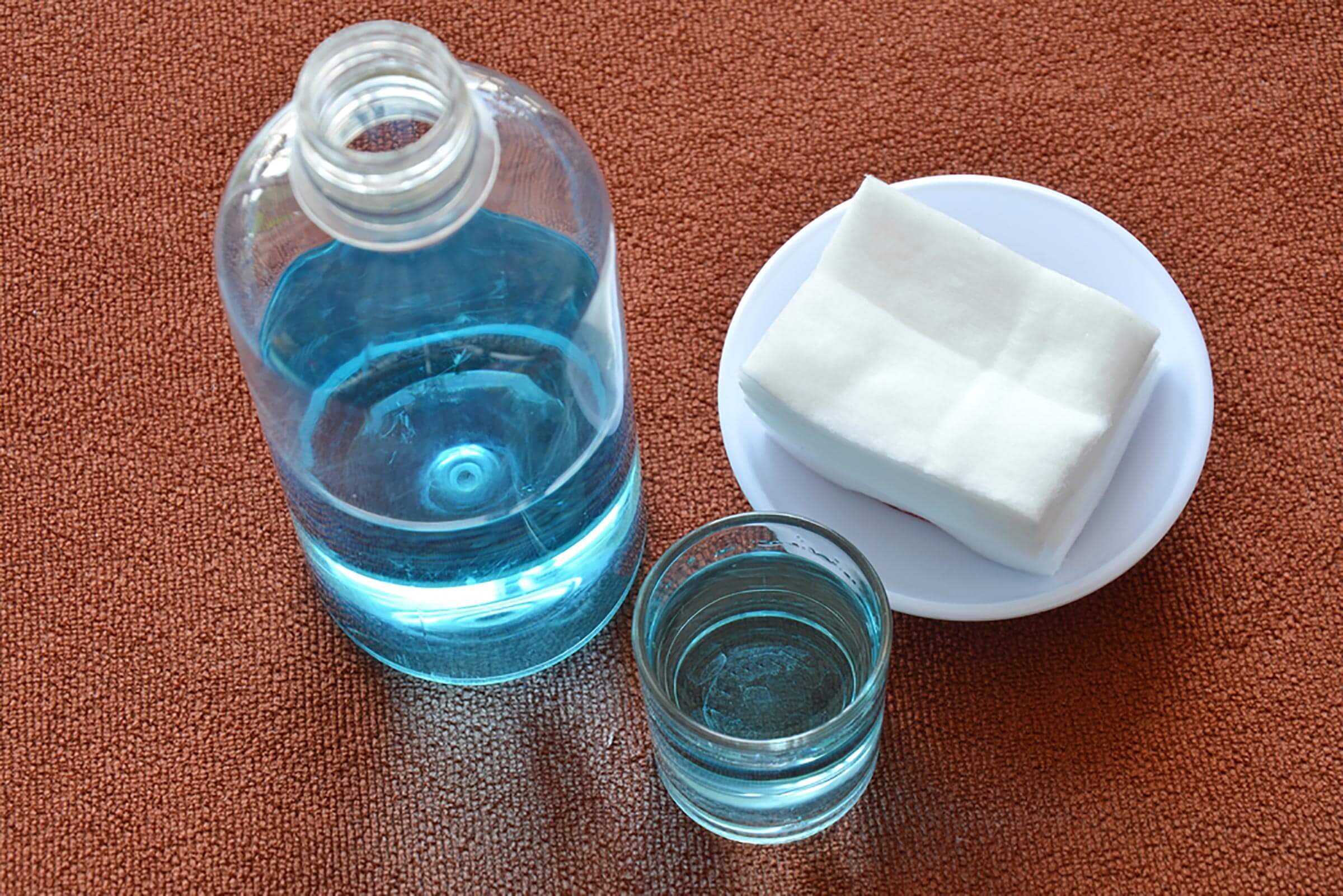 A glass of blue liquid sits beside a bottle of the same liquid, accompanied by a white cloth on a round plate, all on a textured brown surface.