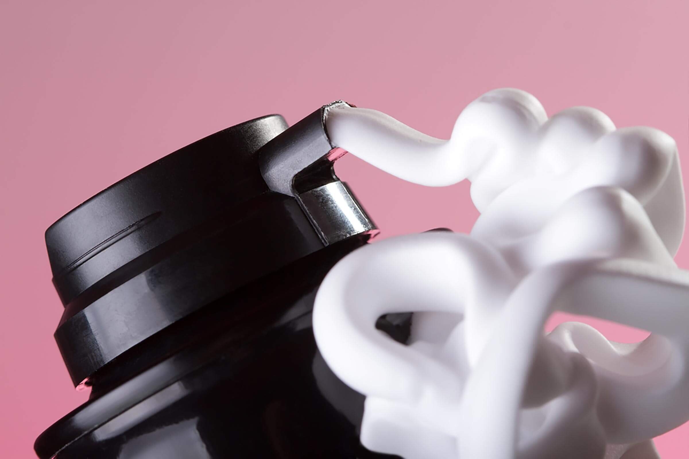 A black bottle cap is slightly ajar, with white foam spilling out. The background is a soft pink, enhancing the bottle's colors.