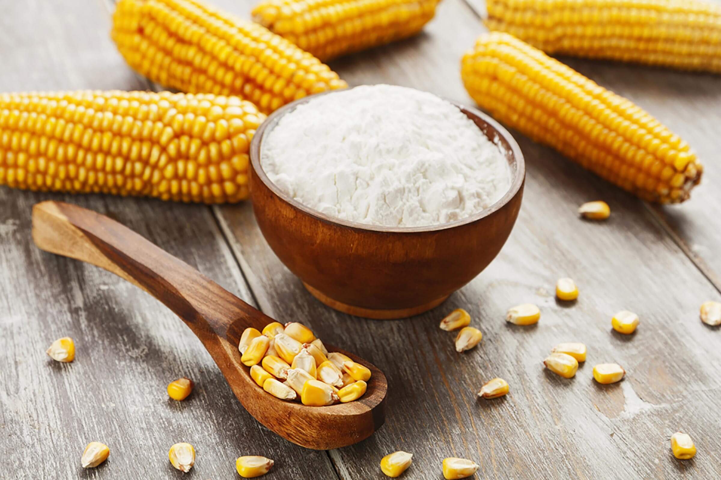 A wooden bowl holds white flour, while yellow corn kernels scatter nearby, accompanied by several ears of corn on a textured wooden surface.