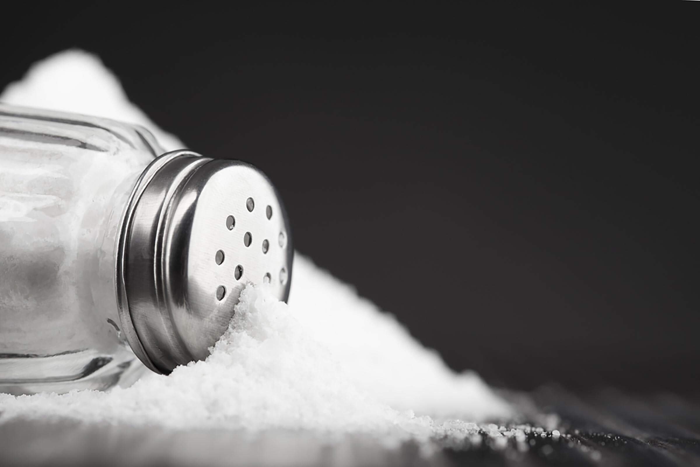 A salt shaker lies on its side, spilling salt onto a dark surface, creating a white mound against a black background.