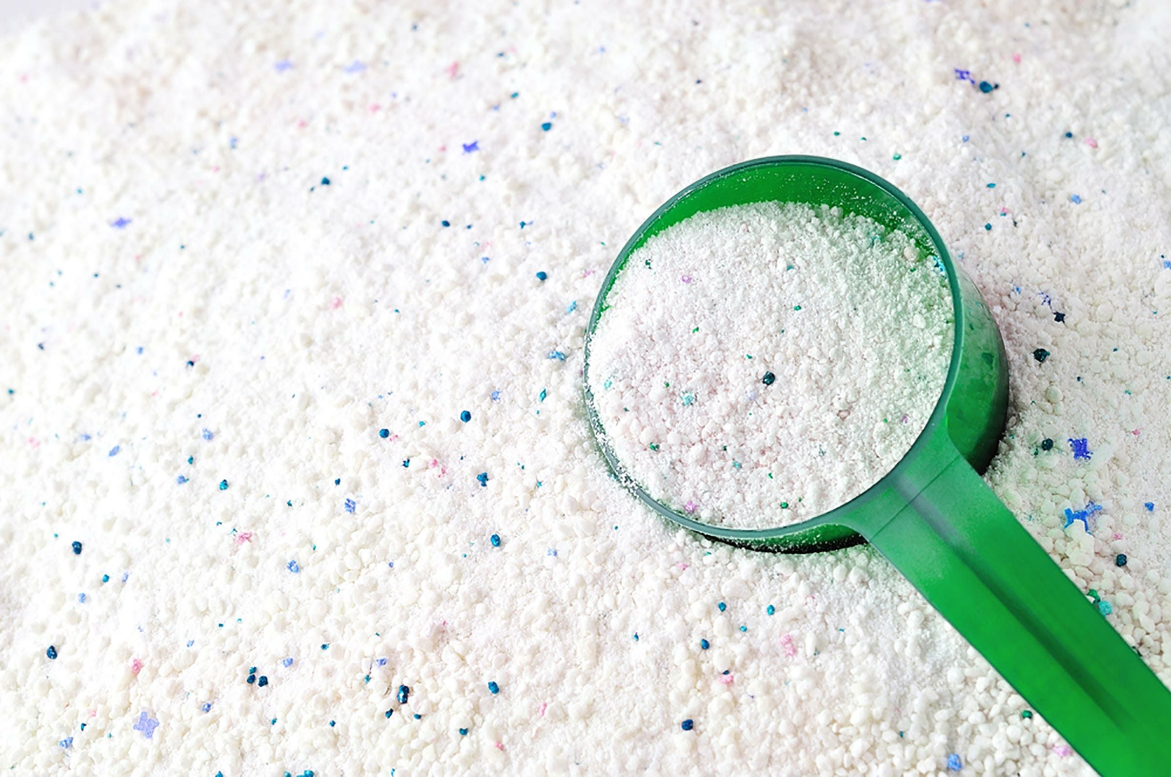A green scoop rests on white powder sprinkled with colorful specks, indicating a laundry detergent on a flat surface.