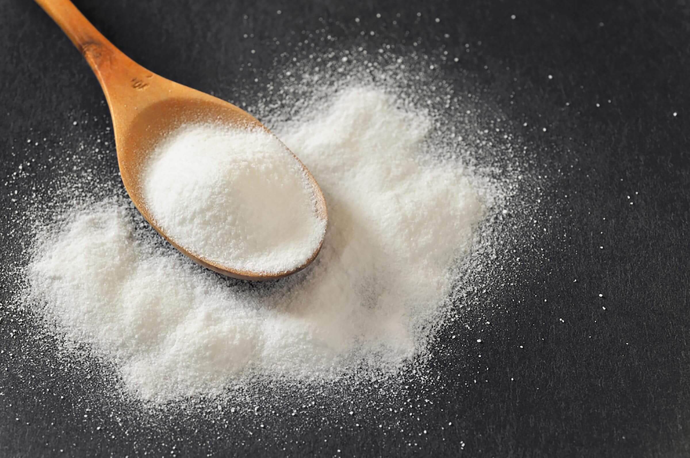 A wooden spoon holds a mound of white powder, surrounded by additional scattered powder, resting on a dark surface.