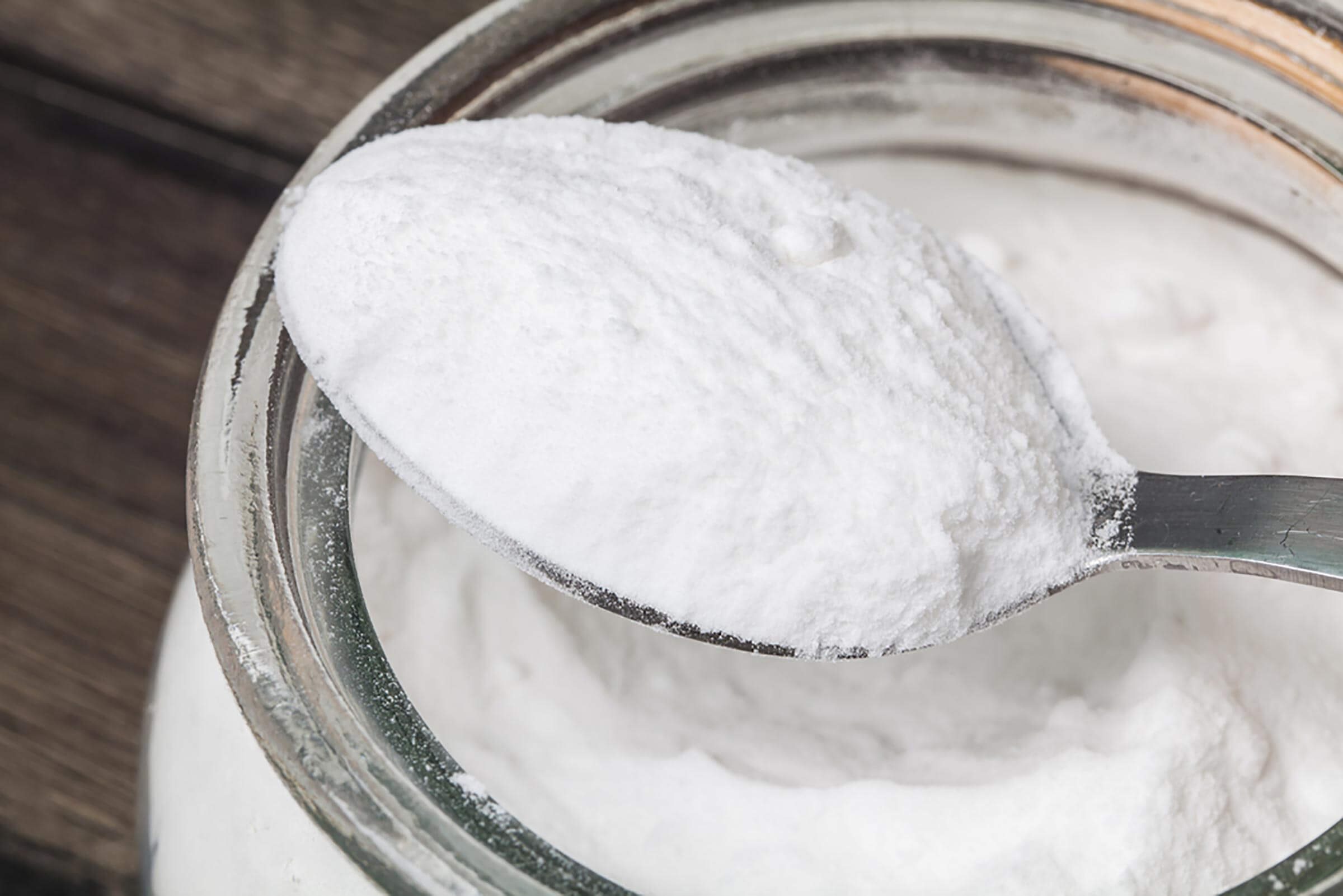 A spoonful of white powder is lifted from a glass jar, resting against a textured wooden surface, suggesting a kitchen setting.