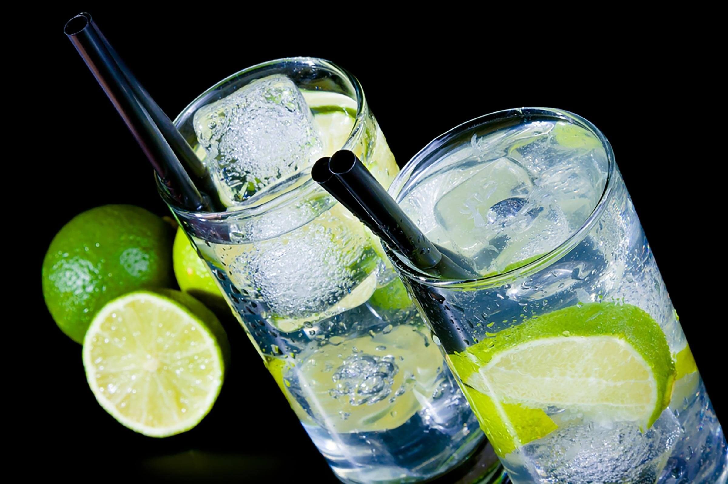 Two tall glasses filled with ice water and lime slices are positioned near whole limes, against a black background. Straws extend into each glass.