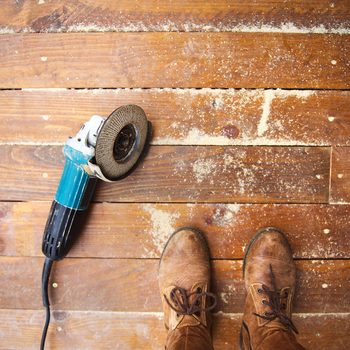 A power tool lies on wooden floorboards, dust scattered around, while a pair of brown boots stands nearby, suggesting ongoing work.