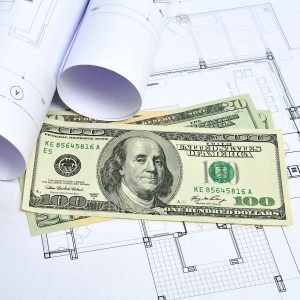Cash bills rest on architectural blueprints, with rolled plans partially visible, indicating a connection between finance and design.