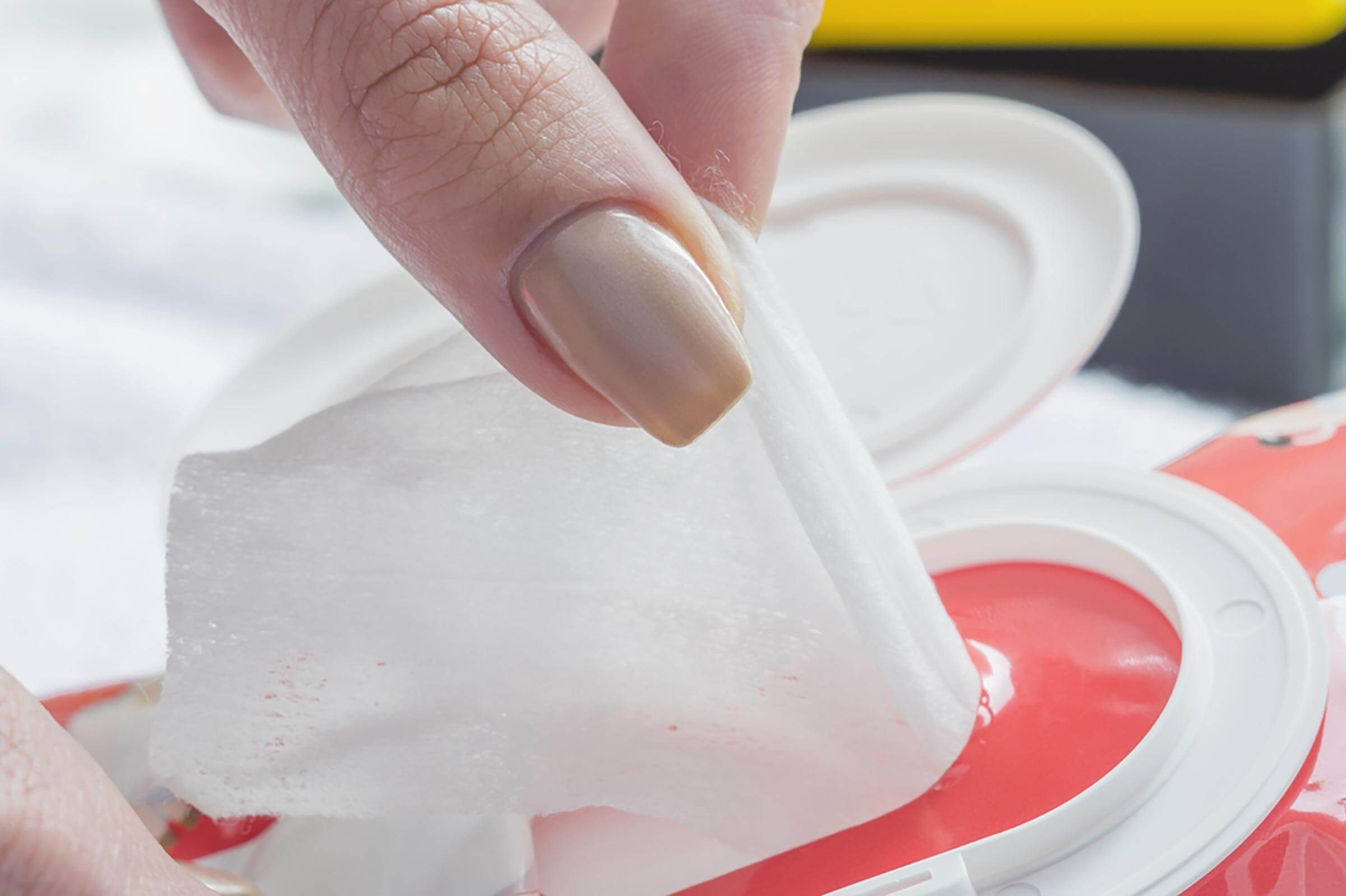 A person pulls a cleansing wipe from a red container, revealing its textured surface against a softly blurred light background.