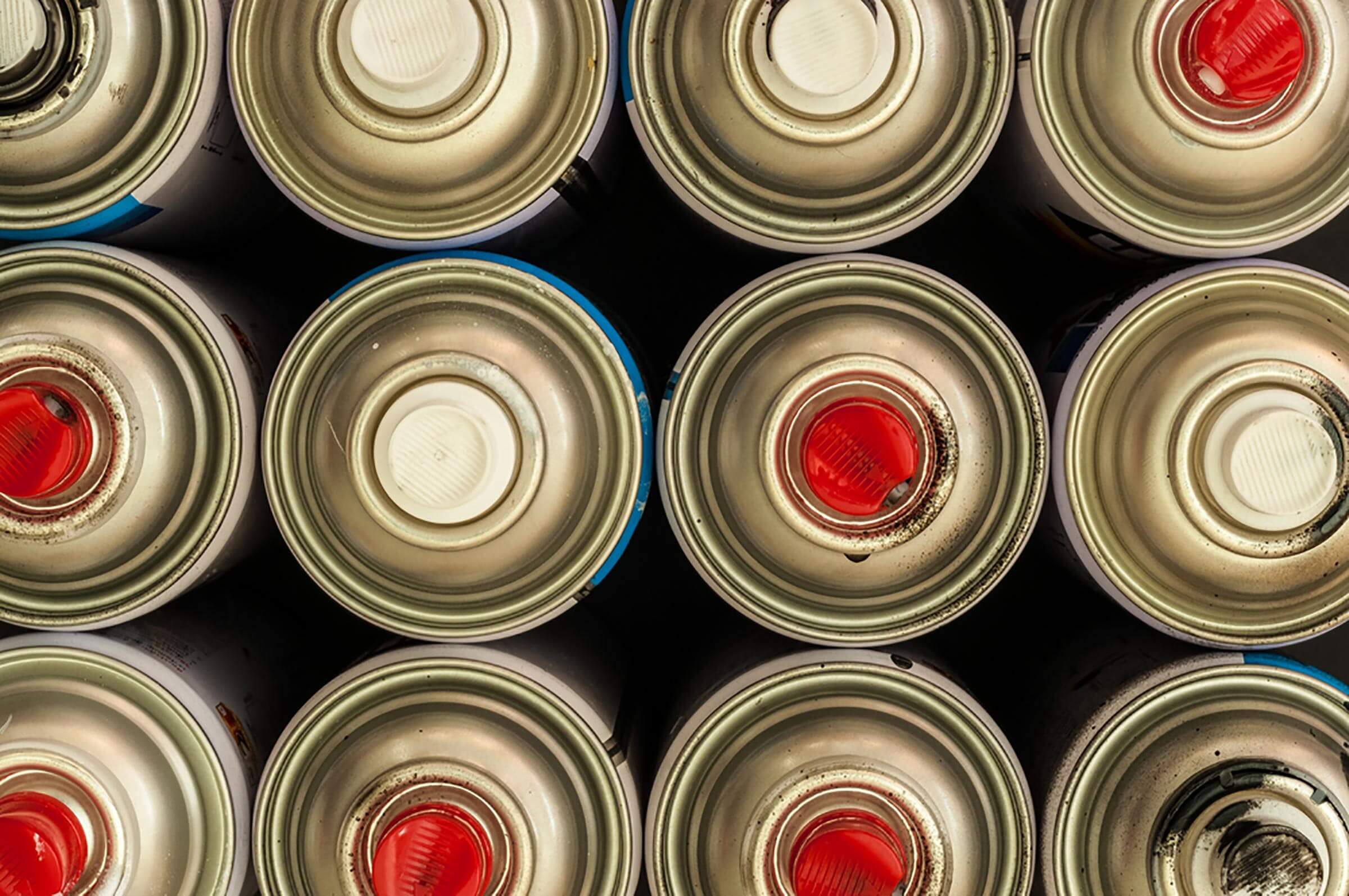 Cans are arranged closely, with some having red and white caps, against a dark background, suggesting they contain spray paint or similar products.