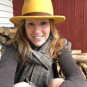A woman with a yellow hat smiles while sitting against a stack of logs, wearing a scarf and a sweater, with a red wooden wall behind her.