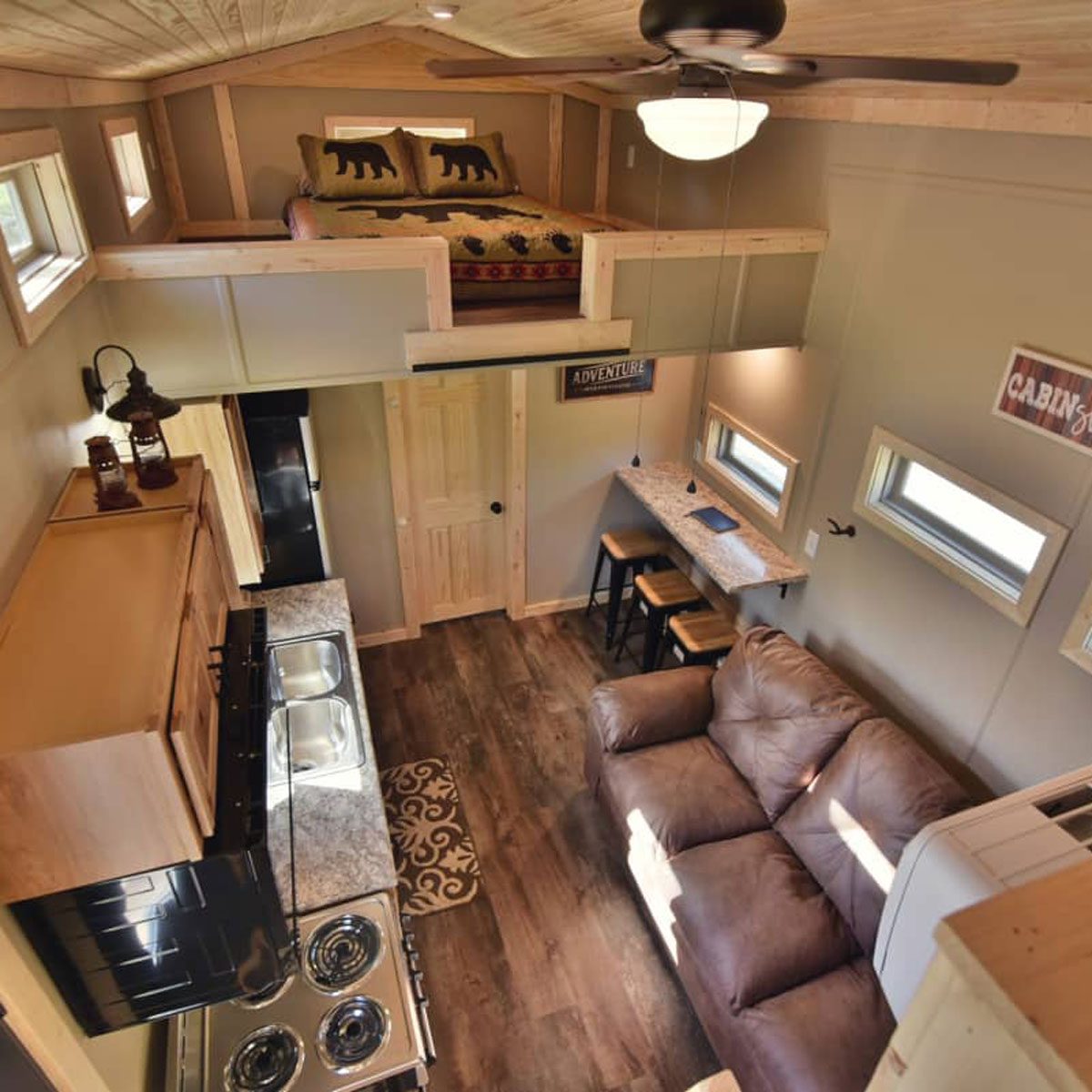 A cozy tiny house features a lofted bed, a kitchen with appliances, a sofa, and a dining area, illuminated by natural light through windows.