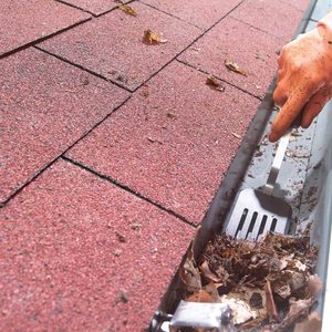 A gloved hand removes leaves from a gutter with a spatula while standing on a sloped red shingle roof under bright lighting.