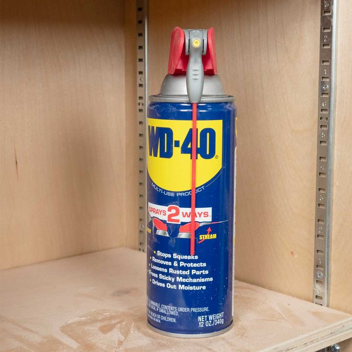 A blue WD-40 spray can stands upright on a wooden shelf, featuring a red nozzle and cap, surrounded by a clean, light wooden backdrop.