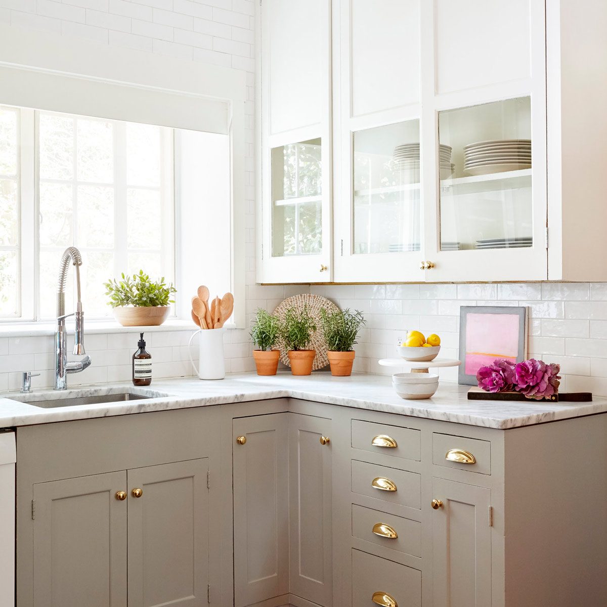 A kitchen features a marble countertop, a sink, potted herbs, wooden utensils, and decorative items, illuminated by natural light through a window.