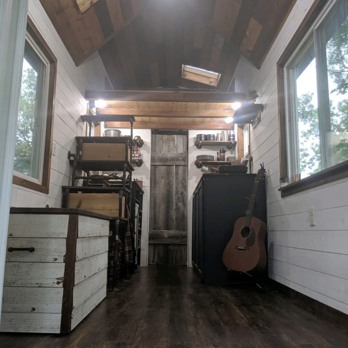 A wooden guitar leans against a cabinet inside a bright, cozy cabin, featuring rustic decor, shelving, and a door at the far end.