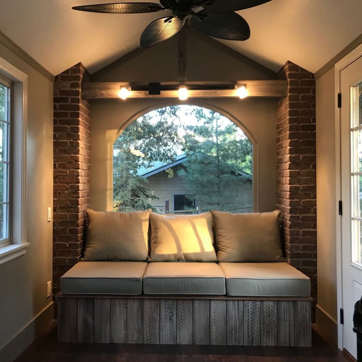 A cozy cushioned bench sits beneath a light fixture, framed by brick walls and a large arched window, offering a view of a wooded area outside.