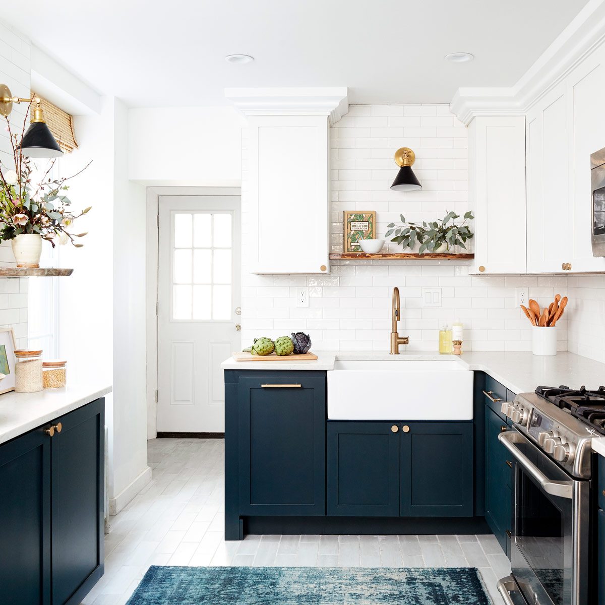 A stylish kitchen features a white backsplash, navy cabinets, a farmhouse sink with fresh produce, and gold accents, illuminated by wall-mounted lamps.