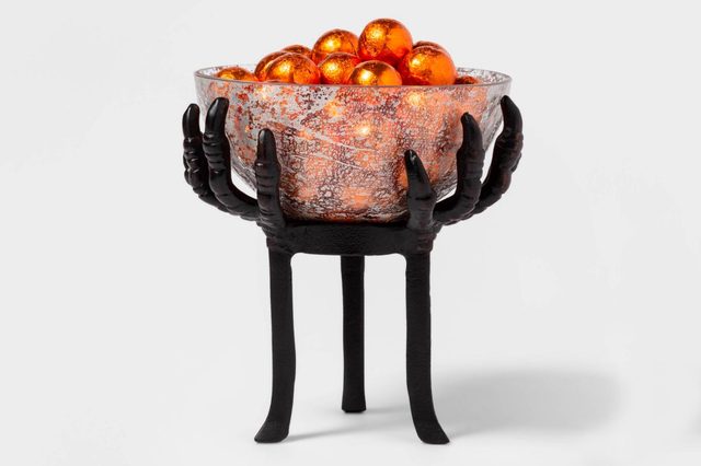 A decorative bowl held by skeletal hands displays shiny orange spheres, set against a plain white background.