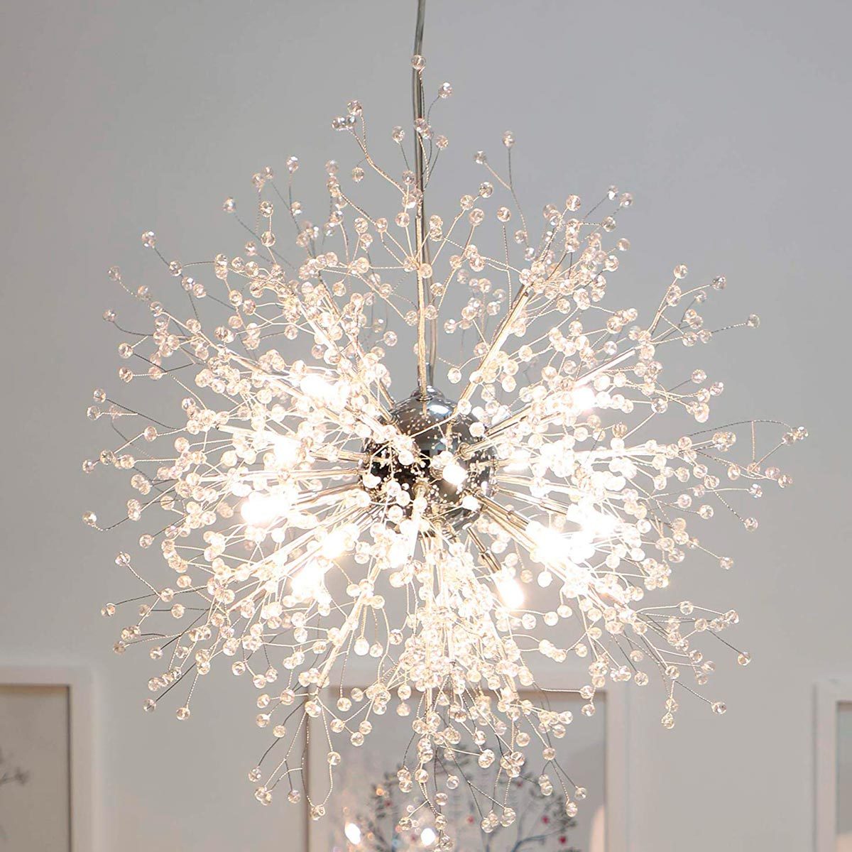A modern chandelier, composed of glass beads and metal, emits bright light, suspended from a ceiling in a minimalist room with framed artwork.