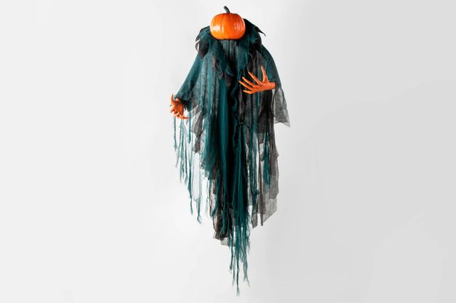 A floating, masked figure with a pumpkin head extends orange hands, draped in long, tattered black and green fabric against a plain white background.
