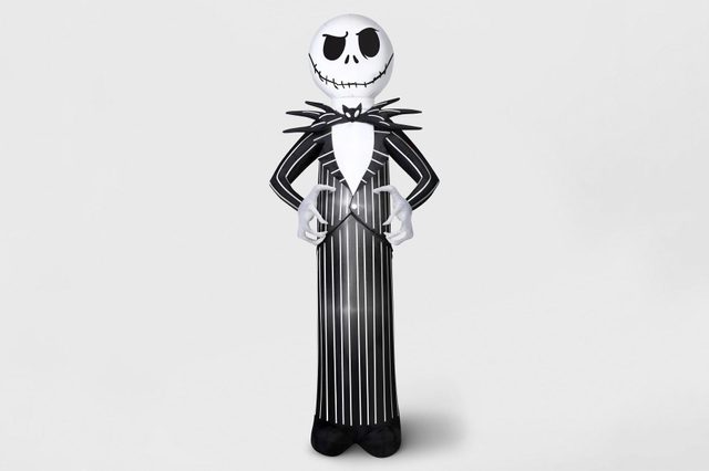 A tall, skeletal figure in a pinstripe suit stands with hands positioned in front, set against a plain, light background.