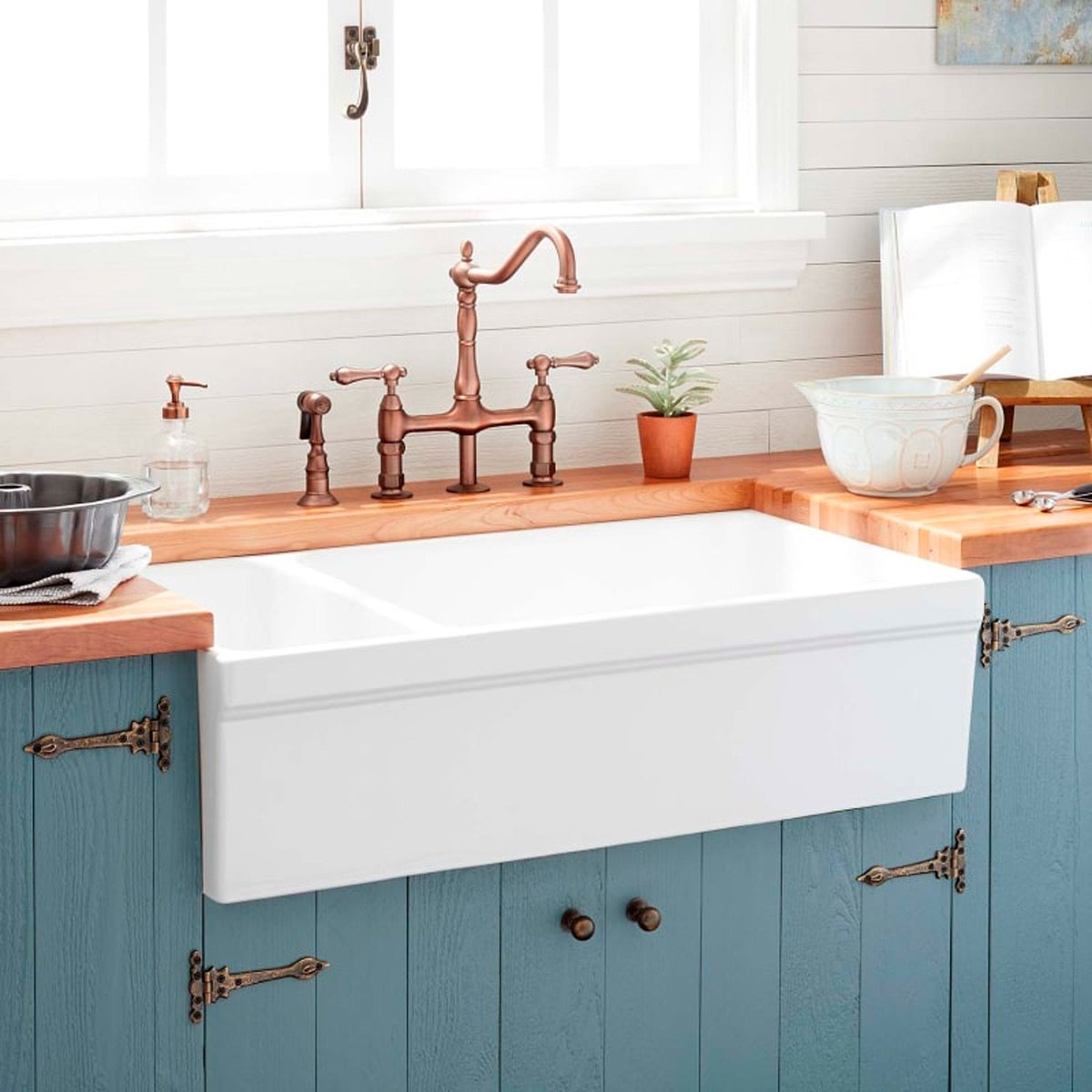 A large white farmhouse sink sits on a wooden countertop, with a bronze faucet above it, surrounded by kitchen utensils and a small potted plant.