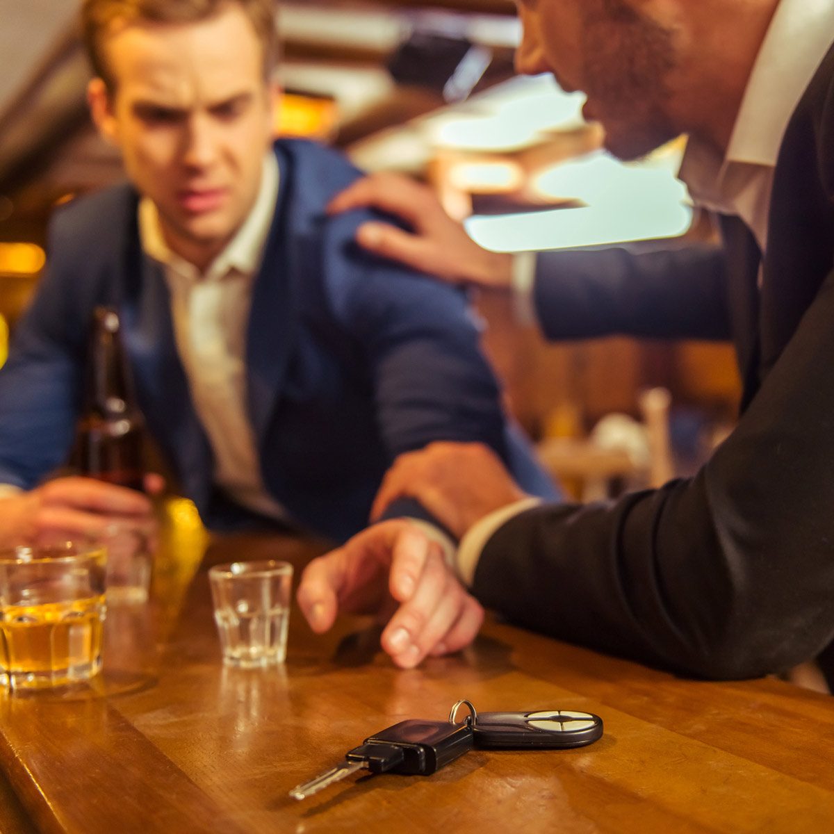 A car key rests on a bar counter as one man reaches for it while another, visibly concerned, places a hand on his shoulder amidst drinks.