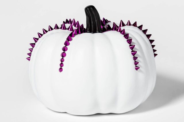 A white pumpkin features a black stem and is adorned with shiny purple spikes, creating a decorative, festive appearance against a neutral background.