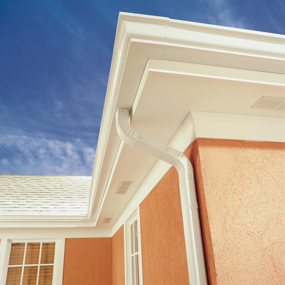 A white gutter channels water along the roof's edge of an orange building, beneath a clear blue sky. Light reflects off the surfaces.