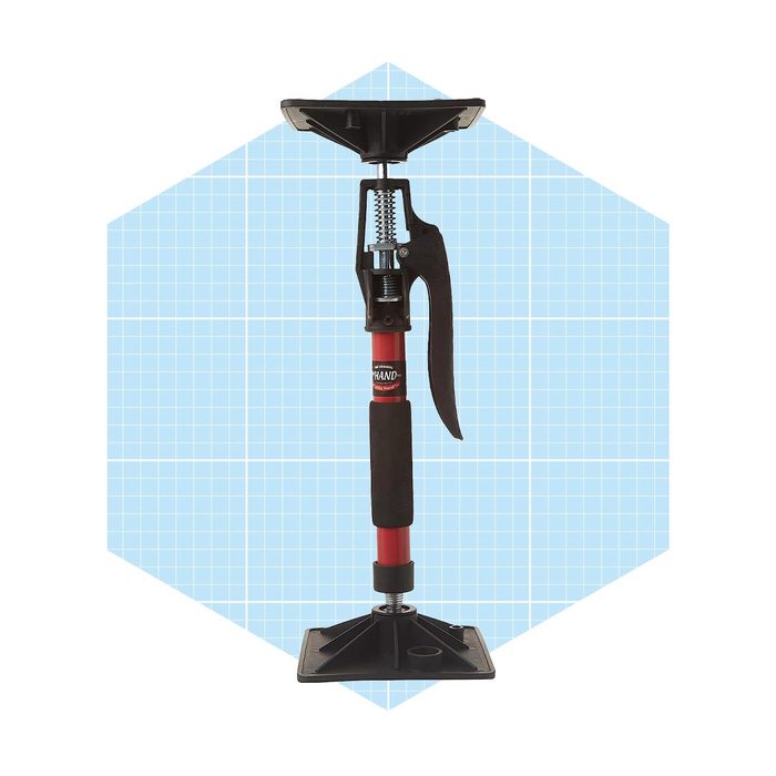 A metal and plastic jack prop stands vertically, with a rubberized grip and base, designed to support weight in construction or repair tasks against a light blue grid background.