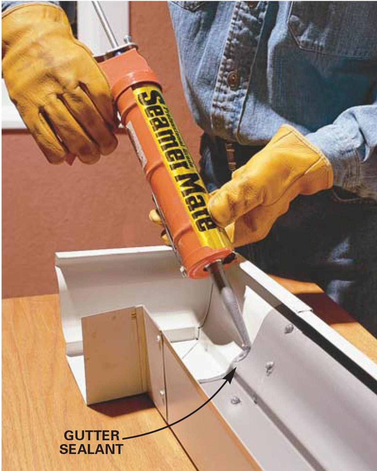 A person in gloves uses a caulking gun to apply gutter sealant into a metal gutter section on a wooden surface.