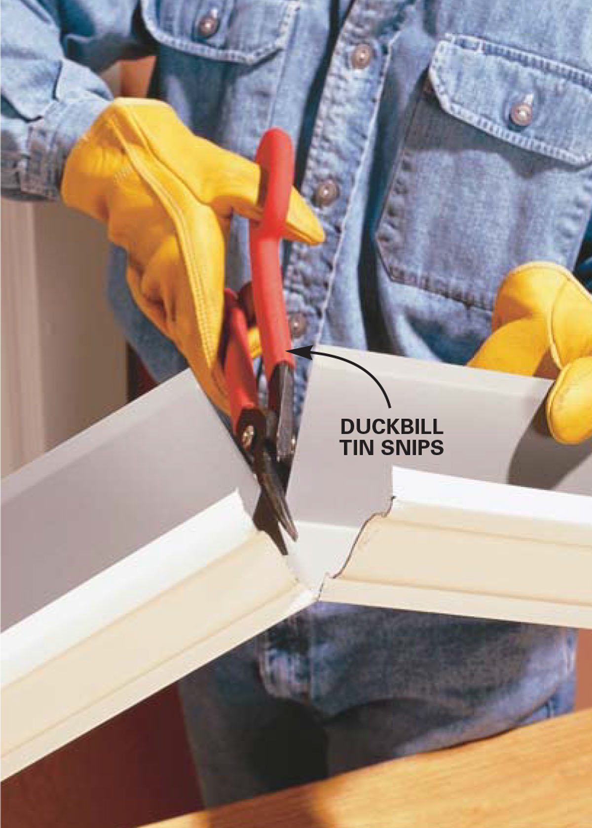 A person wearing yellow gloves uses red tin snips to cut a metal piece in a home environment.