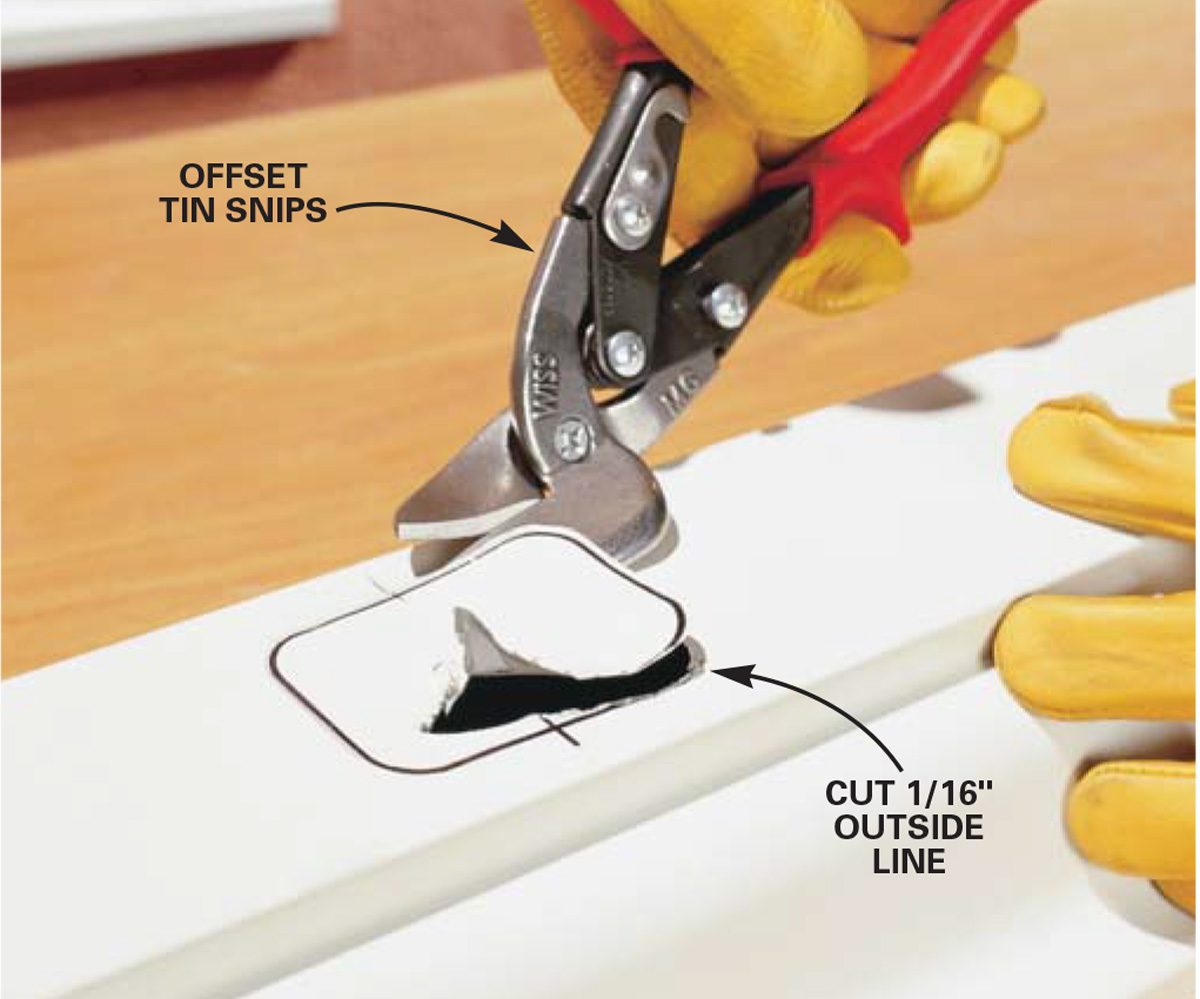 A person uses offset tin snips to cut along a marked line on a white sheet, surrounded by a wooden surface and wearing yellow gloves.