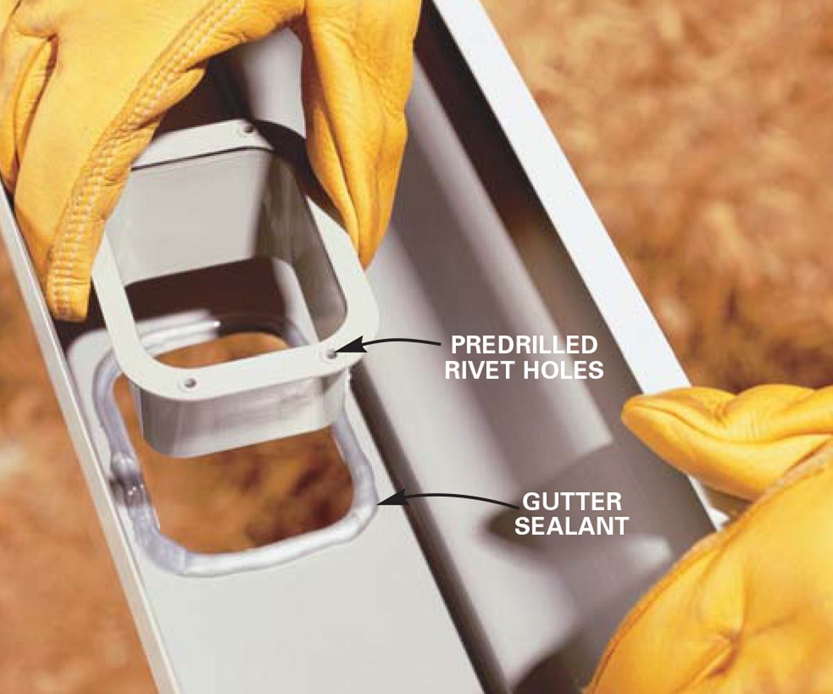 A hand in a yellow glove holds a gutter component, showing rivet holes and sealant, against a textured, earthy background.