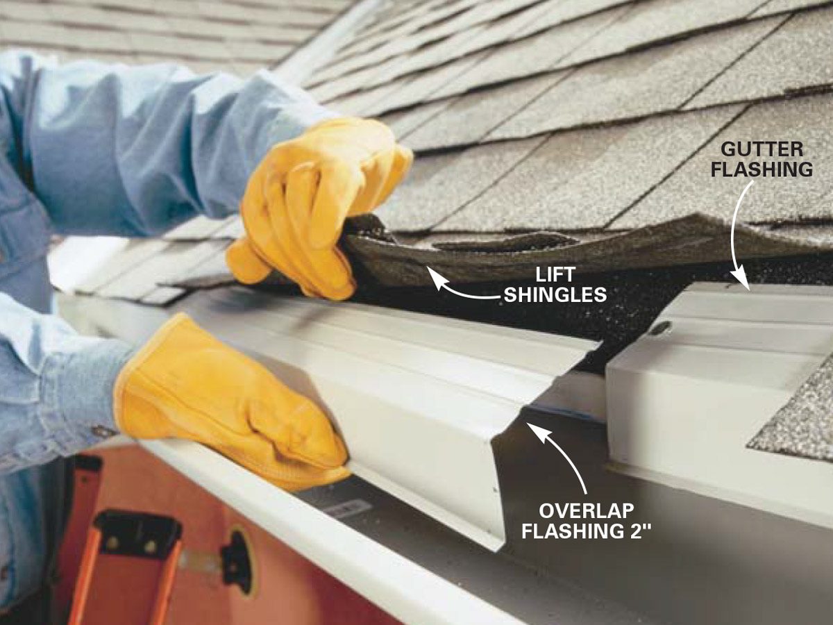A hand in a yellow glove lifts shingles, exposing gutter flashing on a roof under construction, with a ladder visible in the background.