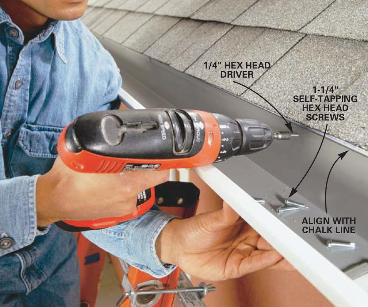A person is using a power drill to install self-tapping screws into a gutter system on a roof, with shingles in the background.