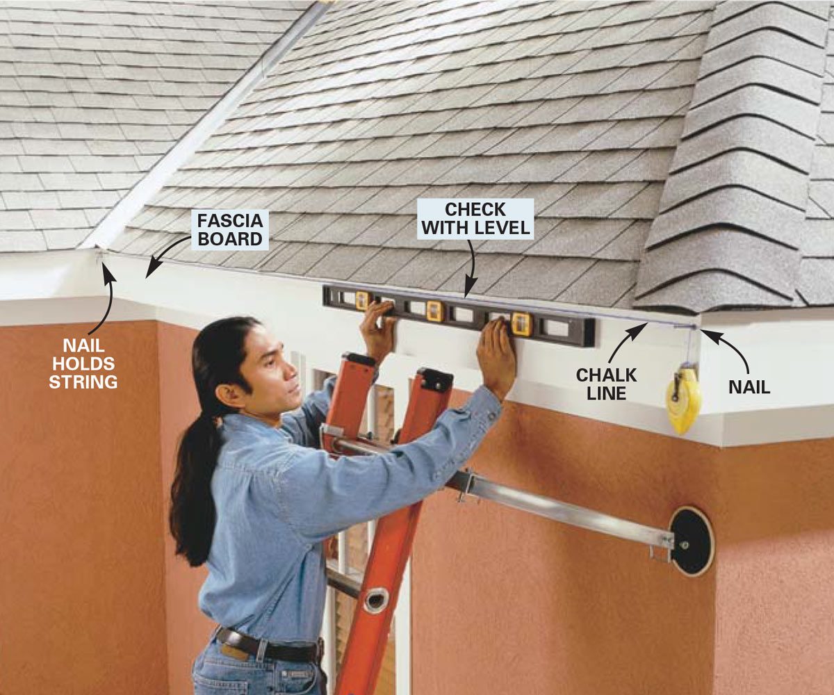 A person uses a level to align a fascia board while standing on a ladder near a sloped roof, with tools and a chalk line visible.