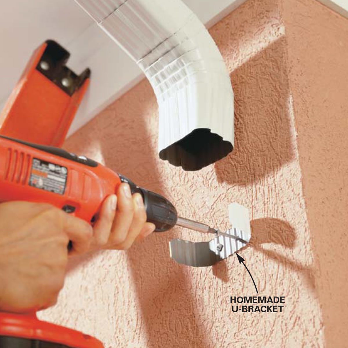 A person drills a wall, securing a homemade U-bracket to support a white, curved gutter on a textured pink surface.