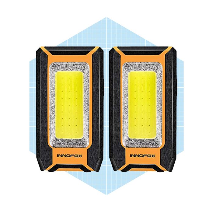 Two portable work lights are displayed, illuminating their yellow LED surfaces, set against a light blue geometric background.