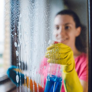 A person in yellow gloves sprays window cleaner on a foggy glass surface while smiling, surrounded by a softly lit indoor environment.