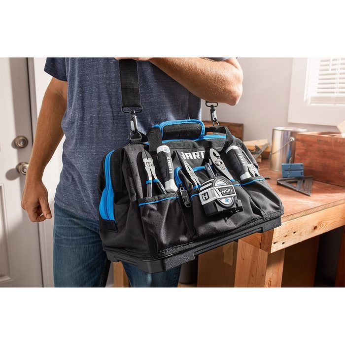 A person holds a tool bag filled with various tools, standing in a well-lit room near wooden furniture and items like a paint can and square.