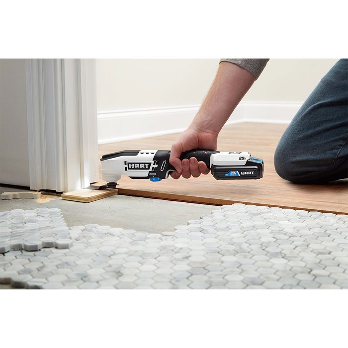 A person uses a powered cutting tool to trim flooring near a doorframe, with hexagonal tiles laid out nearby on a concrete surface.