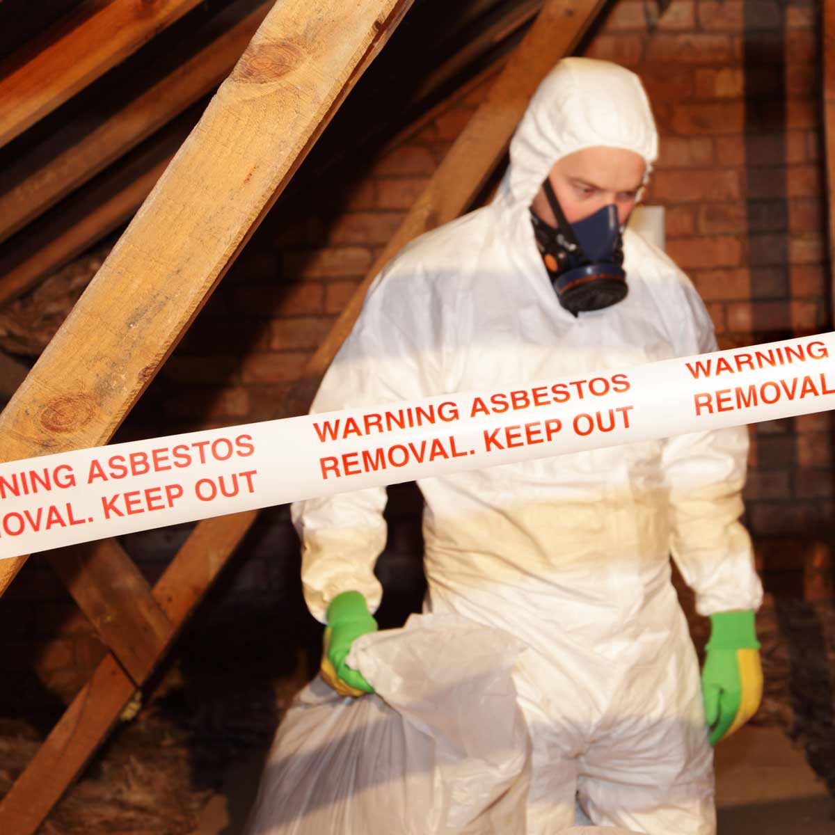 A person in a protective suit and mask removes waste in an attic, while surrounded by "WARNING ASBESTOS REMOVAL. KEEP OUT" tape.