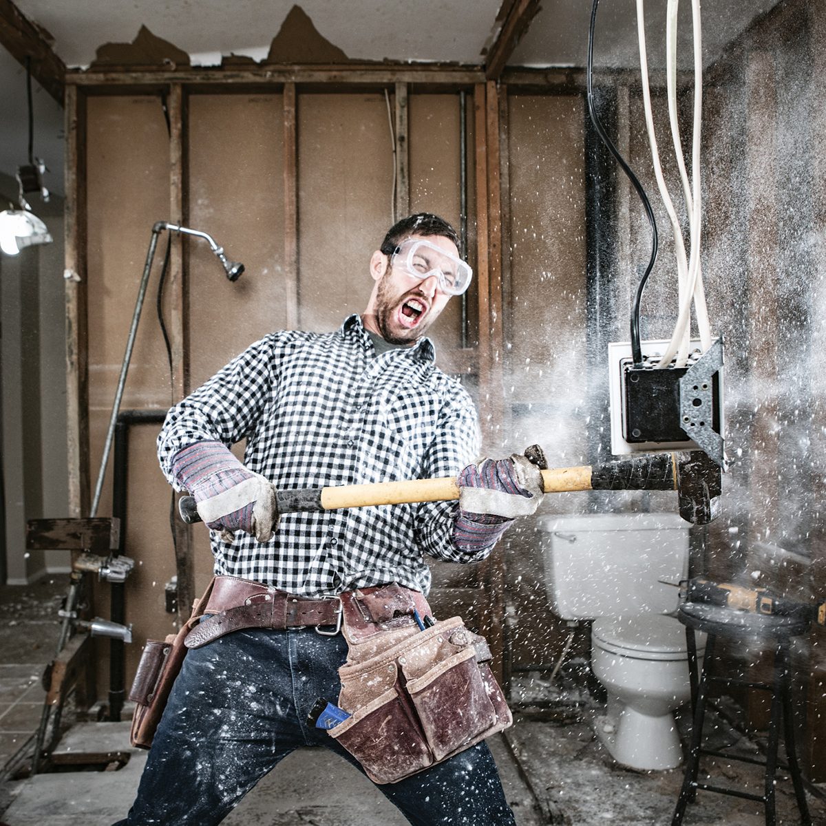 A man swings a sledgehammer, creating debris, in a partially constructed room with plumbing fixtures and exposed wiring in the background.