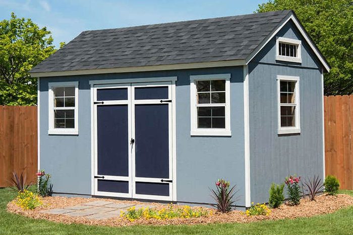 A blue shed with a gray roof stands in a landscaped yard, featuring flowers and shrubs, against a backdrop of green trees and a wooden fence.