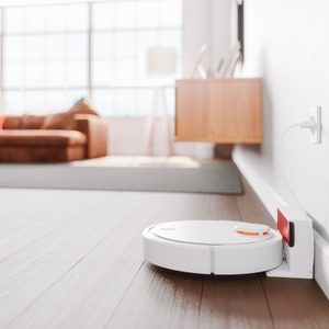 A robotic vacuum is docked against a wall, charging. Sunlight illuminates a spacious, modern living room with a couch and a decorative lamp.