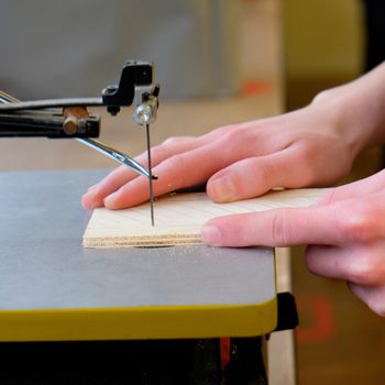 A person's hands guide a piece of wood against a saw blade, cutting through the material on a workbench in a workshop setting.