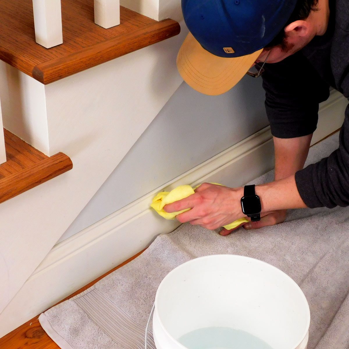 A person cleans a wall with a yellow cloth while kneeling on a towel, near a bucket of water, beside wooden stairs.